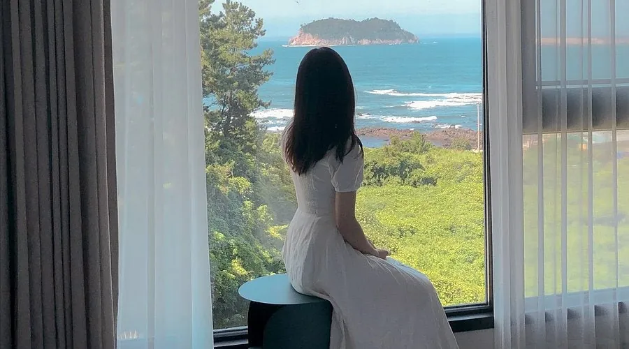 Sitting by a large window with sheer curtains, looking out at the sea and an island